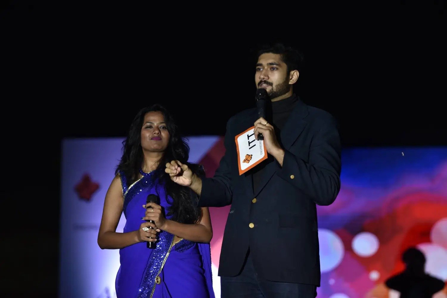 Man and woman speaking on stage at night with blue lighting